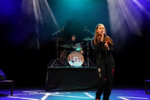 Sixth Form girl singing into a microphone on stage with a drummer in the background